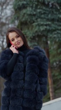 Doğanın arka planında kalın paltoyla güzel bir model. Kürklü, kırmızı telefonla konuşan ve kış parkında kameraya bakan güzel bir kızın portresi. Dikey video