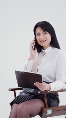 Stüdyoda izole bir şekilde telefonla konuşan esmer kız. Beyaz bluzlu genç bir iş kadını elinde dosyayla sandalyede oturuyor ve cep telefonuyla konuşuyor. Ağır çekim. Dikey video