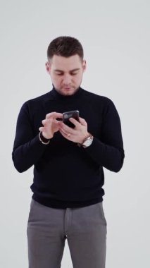 Cep telefonu olan yakışıklı bir adam stüdyoda izole edilmiş. Telefonuna bakan siyah kazaklı ve gri pantolonlu bir gencin portresi. Dikey video