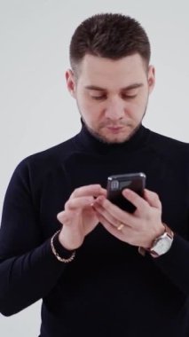 Konsantre genç adam telefona bakıyor. Modern bir akıllı telefon tutan siyah kazaklı yakışıklı bir adam stüdyoda izole edilmiş bir şekilde ona bakıyor. Dikey video