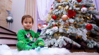 Yeşil pijamalı güzel bir çocuk Noel ağacının yanında oturuyor. Tatil için evde sevimli bir bebek..