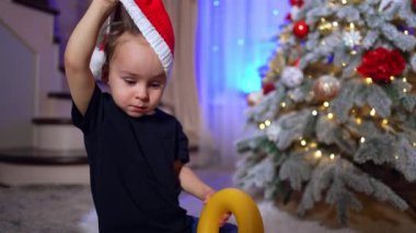 İki yaşındaki çocuk Noel Baba şapkasını çıkarıyor. Komik bebek yüzüne sarı oyuncak yüzük takıyor. Arkaplanda Noel ağacı.