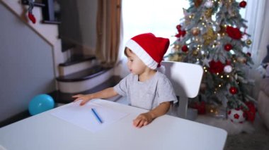 Kırmızı şapkalı sevimli erkek bebek elinde kalemle masadan kalkar. Çocuk masanın arkasına saklanarak yere oturuyor..