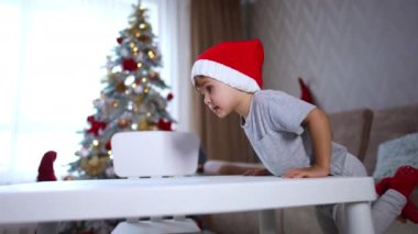Beyaz, gri pijamalı, kırmızı Noel Baba şapkalı çocuk masanın yanında duruyor. Çocuk masaya eğiliyor ve ayaklarını kaldırıyor. Arkaplanda bulanık bir Noel ağacı.