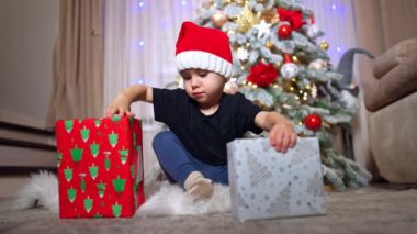 Ciddi bir beyaz çocuk hediye kutularını yönetiyor. Kırmızı Noel Baba şapkalı çocuk Noel ağacının yanında oturmuş hediyelerle uğraşıyor..