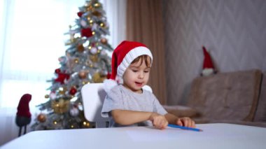 Masada oturan kırmızı şapkalı güzel erkek bebek. Mavi kalemle yeni yürümeye başlayan çocuk çiziyor. Arkaplanda bulanık bir Noel ağacı.
