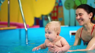 Sevimli tombul oğlan havuza su sıçratıyor. Kadın kucağında bir çocukla ona yüzmeyi öğretiyor..