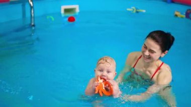 Pozitif kadın bebeğini havuza taşıyor. Küçük bebek bir oyuncak çiğniyor. Bir çocuğa doğduğundan beri yüzmeyi öğretmek..
