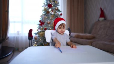 Mavi kalemle kırmızı Noel Baba şapkası çizen odaklanmış bebek. Yeni yürümeye başlayan çocuk Noel tatilinde.