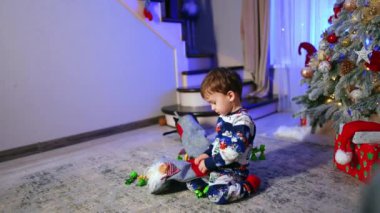 Kışın pijamalı siyah saçlı erkek bebek Noel ağacının yanında oturuyor. Tatlı çocuk çorabından şeker alıyor..