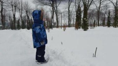 Mavi elbiseli beyaz çocuk kışın ayakta duruyor. Yan görüş. Küçük çocuk önündeki bir şeyi işaret ediyor..