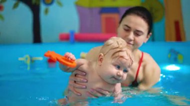 Pozitif esmer kadın çocuğu suya taşıyor. Sevimli bebek oyuncağı çiğniyor. Bebekler için yüzme dersi.