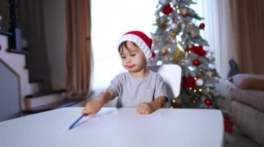 Kırmızı Noel Baba şapkalı, kurşun kalemle çizen beyaz bir bebek. Küçük çocuk resmine parmağıyla işaret ediyor. Arkaplanda Noel ağacı.