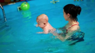 Esmer kadın, yüzme havuzunda bebek tutuyor. Sevimli çocuk suda yüzmek için ellerini hareket ettiriyor..