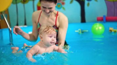 Kadın koç yüzme havuzunda elinde tombul bir çocuk tutuyor. Küçük bebek ellerini sertçe çırpıyor..