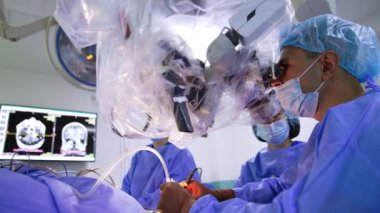 Dikkatli beyin cerrahı modern teknolojik mikroskop kullanarak beyin ameliyatı yapıyor. İki kadın hemşire ameliyat odasında cerraha yardım ediyor. Düşük açı görünümü.