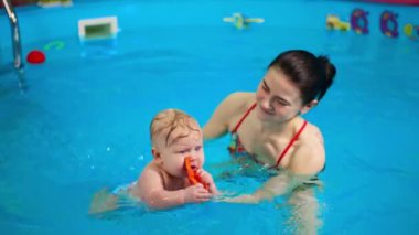 Gülümseyen esmer kadın, bebeği havuza taşıyor. Küçük çocuk suya su sıçratır neşeyle yüzmeyi öğrenir..