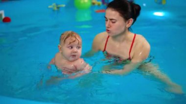 Dolgun beyaz bebek yüzme havuzuna taşınıyor. Bayan antrenör bebeğe yüzmeyi öğretiyor..