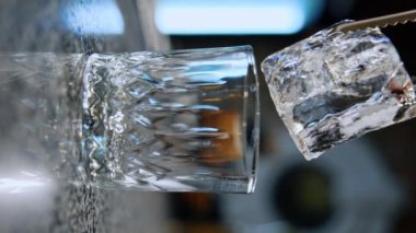 Büyük buz küpü cımbız kullanılarak bir bardağa konur. Barda içkiyi hazırlıyorum. Kapatın. Dikey ekran. Bulanık arkaplan.