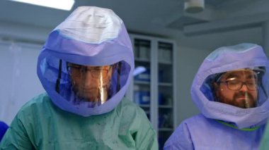 Doktorlar operasyonda koruyucu giysi ve kask giyerler. Cerrahın elbisesi kan lekeleriyle kaplı..