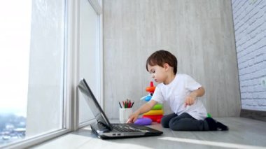 Beyaz tişörtlü beyaz çocuk balkon zemininde oturuyor. Bebek dizüstü bilgisayara bakar ve klavyedeki tuşlara basar..