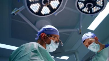 Ameliyat için donanımlı ve üniformalı iki erkek cerrah. Sıhhiyeciler yuvarlak lambalar altında ameliyatta çalışıyor. Düşük açı görünümü.