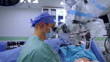 Maskeli orta yaşlı beyaz cerrah mikroskoba dikkatlice bakıyor. Yan görüş. Doktor kullanımda metal araç kullanıyor.
