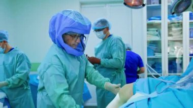 Koruyucu kasklı erkek cerrah hastanın yanında duruyor. Doktor hastanın bacağını bükmeye çalışıyor. Sağlık personeli arka planda.