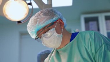 Maskeli, şapkalı ve koruyucu gözlüklü bir erkek doktor. İşteki bir cerrahın yakın portresi..