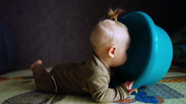 Küçük sarışın bebek önünde bir çamaşır leğeni tutarak yerde yatıyor. Havzayla oynayan sevimli bir çocuk..