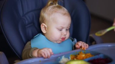 Sevimli beyaz bebek yüksek sandalyede oturuyor. Kadın eli çocuğuna sağlıklı yiyecek verir. Kapat..