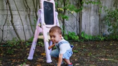 Kot pantolon giymiş yakışıklı beyaz çocuk dışarıda oyuncak arabasıyla oynuyor. Arkaplanda çocuk tahtası.