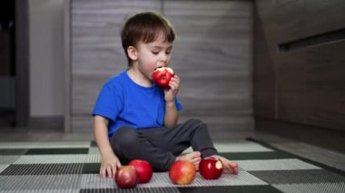 Yerde oturan mavi tişörtlü bebek. Çocuk önündeki birkaç elmadan birini alır, bir ısırık alır ve yerine koyar..