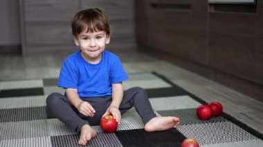 Mutfakta yerde oturan sevimli gülümseyen bebek. Kırmızı elmalar çocuğun yanında yere saçıldı. Çocuk bir meyveyi alıp halıya yuvarlıyor..