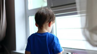 Koyu saçlı erkek bebek pencerede duruyor. Bebek cama bir oyuncak yapıştırır ve kameraya doğru çevirir..