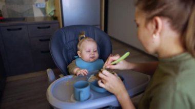 Anne bebeğiyle birlikte yüksek sandalyede oturuyor. Kadın kaşığa biraz avokado alıp çocuğa veriyor..