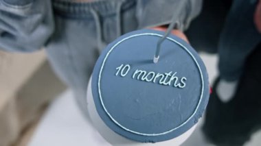 Mavi pasta ve on aylık bir işaret. Pastaya mum yakan tanınmamış kadın eli. Yanında bebekli bir adam duruyor. Üst görünüm.