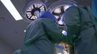 Hastayı mavi cüppeli ve kasklı erkek doktorlar ameliyat ediyor. Arka planda yuvarlak lambalar. Düşük açı görünümü.