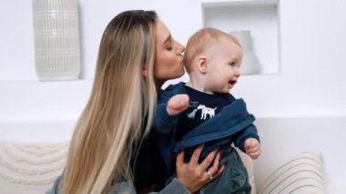 Güzel sarışın, uzun saçlı kadın kanepeye oturup bebeğini tutuyor. Kadın çocuğunu öpüyor ve çocuk itiraz ediyor..