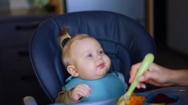 Mavi önlüklü sarışın çocuk yüksek sandalyede oturuyor. Tanınmayan anne çocuğunu kaşıkla besler..