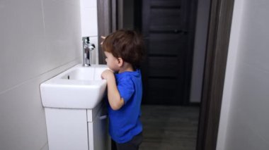 Beyaz çocuk lavabonun yanındaki banyoda duruyor. Meraklı çocuk suyun akışını seyrediyor ve elini suyun altına sokuyor..