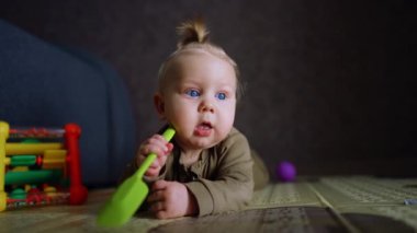 Şirin, tombul yanakları ve mavi gözleri olan küçük bir bebek. Tatlı bebek yerde yatıyor ve oyuncak bir kürek çiğniyor. Kapat..