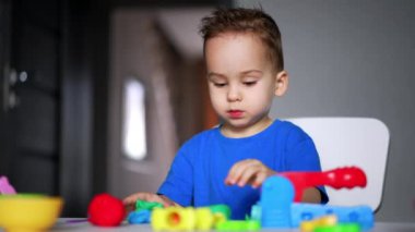 Mavi tişörtlü sevimli erkek bebek oyununa odaklanmış. Bebek plastik bardaklar ve plastikler kullanıyor. Bulanık arkaplan.