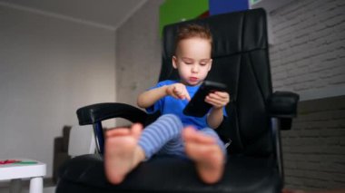 Sevimli beyaz çocuk, ofis sandalyesinde döner koltukta oturur. Tatlı çocuk akıllı telefona odaklanmış durumda. Düşük açı görünümü.