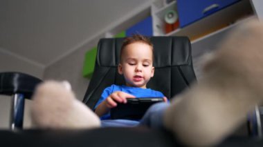 Beyaz çocuk siyah ofis sandalyesinde oturuyor. Ayakları kameraya yakın. Düşük açı elinde akıllı bir telefona bakıyor..