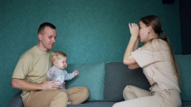 Babamın kucağında ağlayan bebek. Karşıda oturan anne çocuğu eğlendiriyor ve o gülümsüyor..