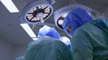 Ameliyat yapan mavi cüppeli ve kasklı iki cerrahın arkası. Aydınlatma ekipmanlarının altındaki sağlık görevlilerinin görüş açısı düşük..