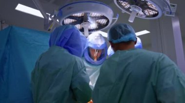 Operasyon masasında yoğun çalışan tıbbi ekibe yaklaşıyoruz. Koruyucu miğferli doktorlar yuvarlak lambalar altında çalışır..