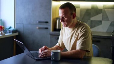 Mutfakta laptopuyla uğraşan Kafkasyalı bir adam. Odaklanmış serbest yazar ekrana bakar ve klavyede yazı yazar.