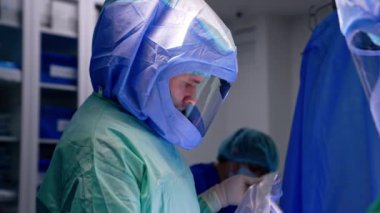 Cüppeli ve kasklı bir erkek doktor elinde bir plastik parçası tutuyor. Yan görüş. Modern cerrahi odasında çalışan sağlık görevlileri..